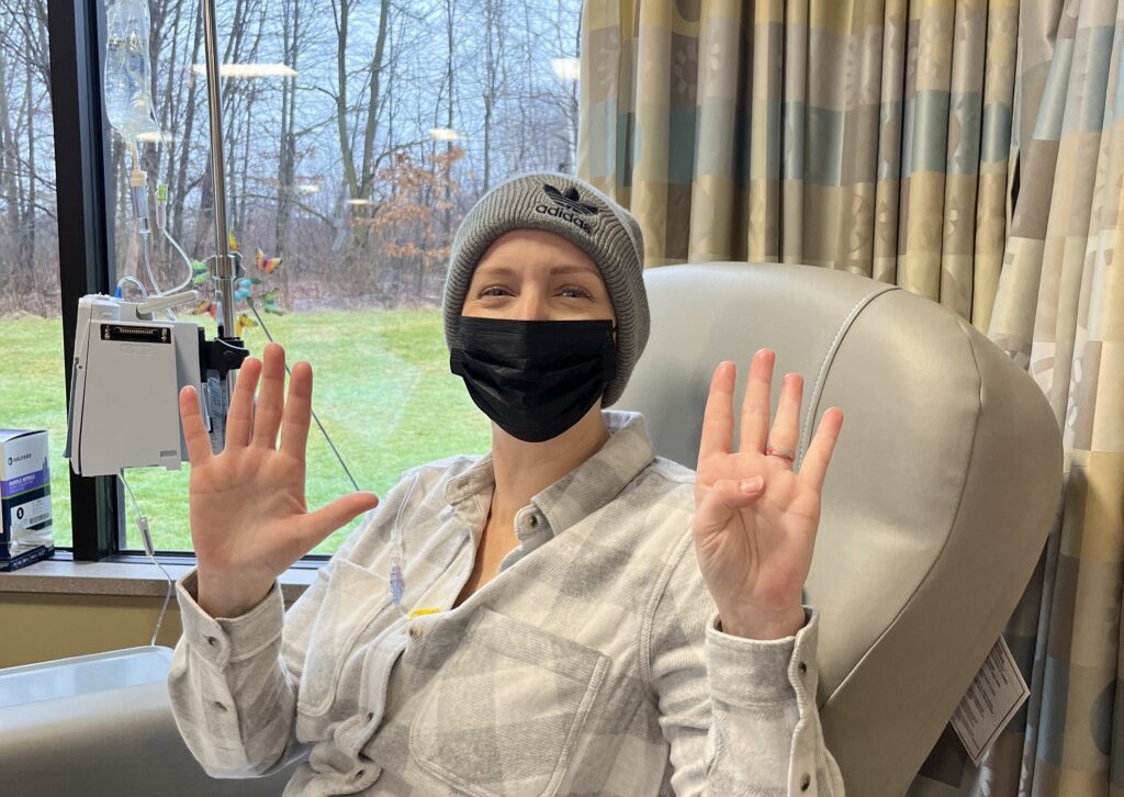 A woman in a hospital chair wears a beanie and black mask, holding up nine fingers -perhaps marking a milestone in their breast cancer journey. IV equipment is visible by the window, with trees and grass outside. The woman is smiling and wearing a plaid shirt.