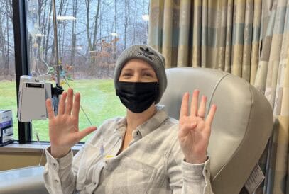 A woman in a hospital chair wears a beanie and black mask, holding up nine fingers -perhaps marking a milestone in their breast cancer journey. IV equipment is visible by the window, with trees and grass outside. The woman is smiling and wearing a plaid shirt.