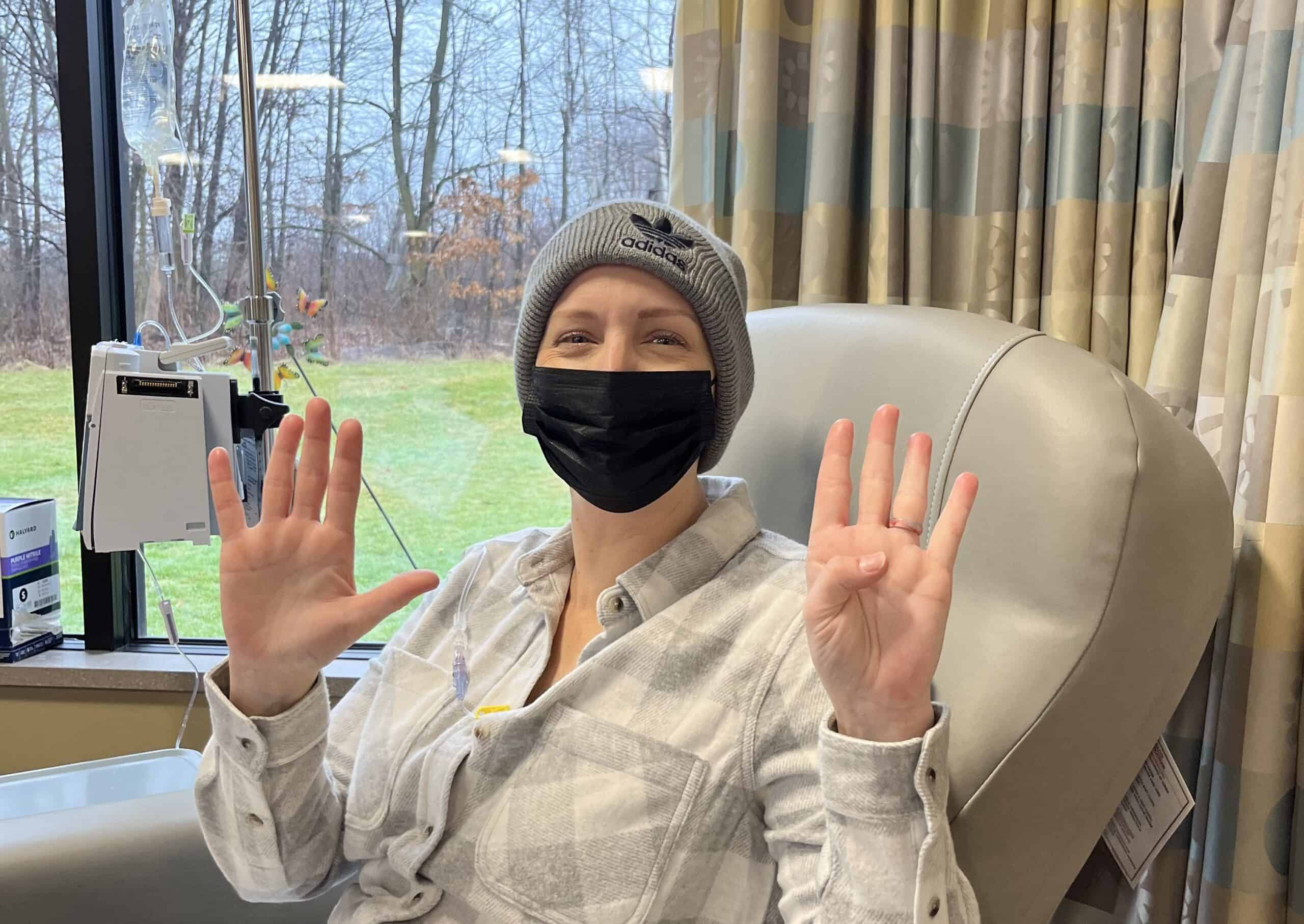 A woman in a hospital chair wears a beanie and black mask, holding up nine fingers -perhaps marking a milestone in their breast cancer journey. IV equipment is visible by the window, with trees and grass outside. The woman is smiling and wearing a plaid shirt.