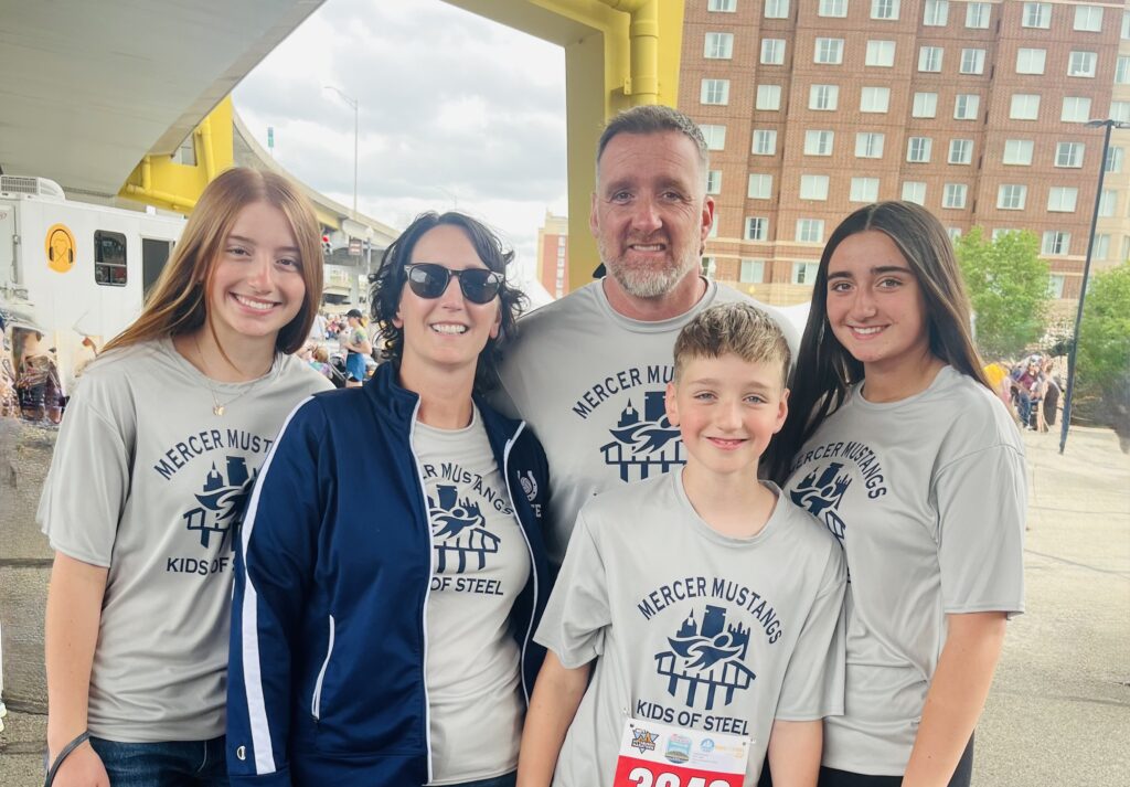 A group of five people wearing matching “Mercer Mustangs Kids of Steel” shirts smiles outdoors at an event, with buildings and a yellow bridge in the background.