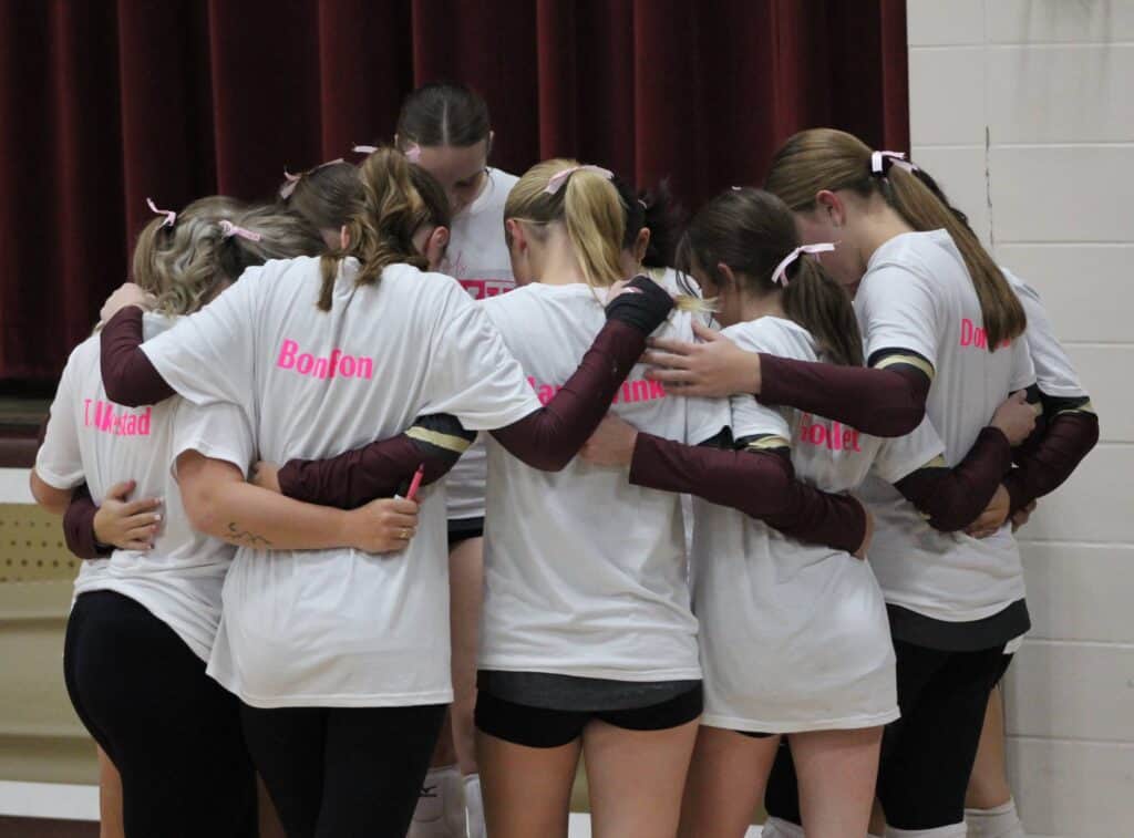 A group of volleyball players in white shirts with pink writing huddle together, arms around each other, preparing for a game during a volleyball breast cancer fundraisers. They are standing in front of a maroon curtain, facing inward.