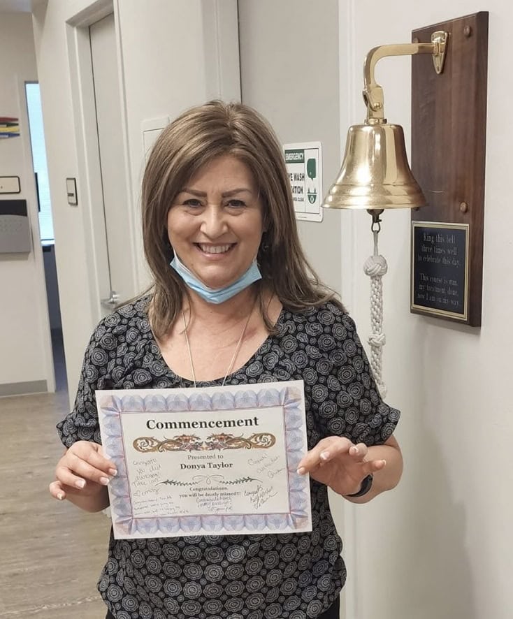 A woman smiles while holding a commencement certificate in recognition of the end of her breast cancer treatment. She stands next to a gold bell mounted on the wall, with her mask pulled down under her chin.