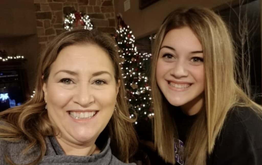 Two women smile in front of a decorated Christmas tree adorned with white lights and ornaments indoors.