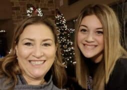 Two women smile in front of a decorated Christmas tree adorned with white lights and ornaments indoors.