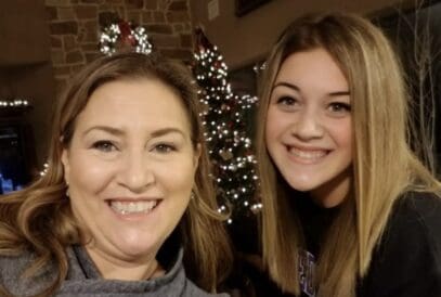 Two women smile in front of a decorated Christmas tree adorned with white lights and ornaments indoors.