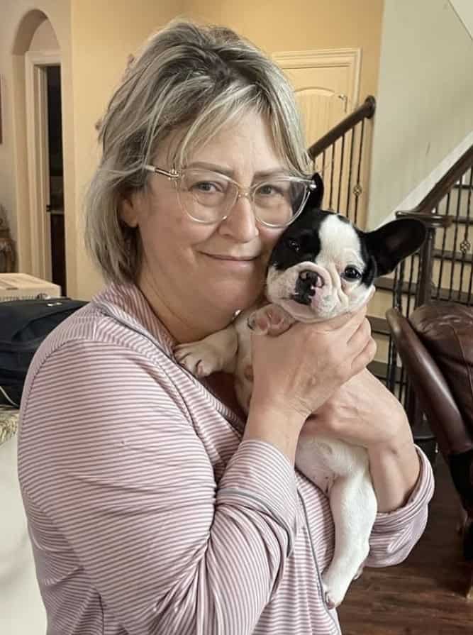 A woman with glasses and short, blonde hair is smiling and holding a black and white French Bulldog puppy indoors near a staircase.