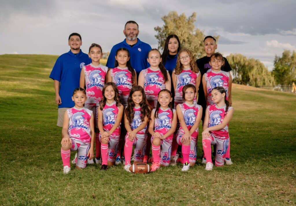 A youth girls’ flag football team in pink uniforms poses outdoors on grass with three coaches standing behind them, smiling for a youth sports breast cancer fundraiser. A football sits in front, with trees and a cloudy sky in the background.