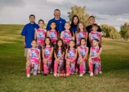 A youth girls’ flag football team in pink uniforms poses outdoors on grass with three coaches standing behind them, smiling for a youth sports breast cancer fundraiser. A football sits in front, with trees and a cloudy sky in the background.