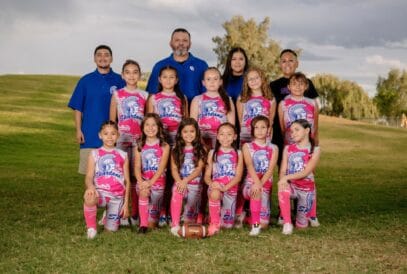 A youth girls’ flag football team in pink uniforms poses outdoors on grass with three coaches standing behind them, smiling for a youth sports breast cancer fundraiser. A football sits in front, with trees and a cloudy sky in the background.