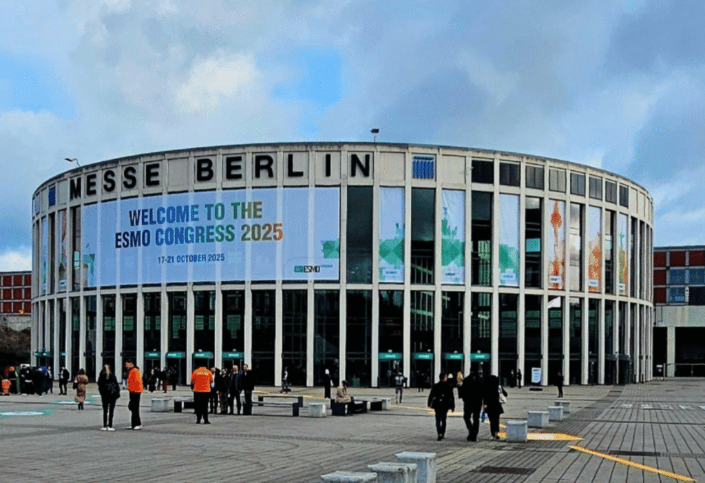 The Messe, Berlin building is pictured with a sign that reads “Welcome to the ESMO Congress 2025” as people walk in front of the building