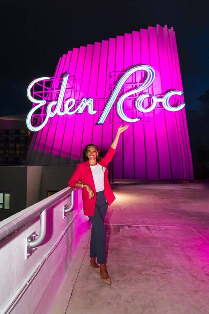 A woman in a red blazer smiles and points up at a large, glowing neon “Eden Roc” sign at night. Illuminated in bright pink, the vibrant background captures the spirited atmosphere as Florida Goes Pink for National Breast Cancer Awareness Month.