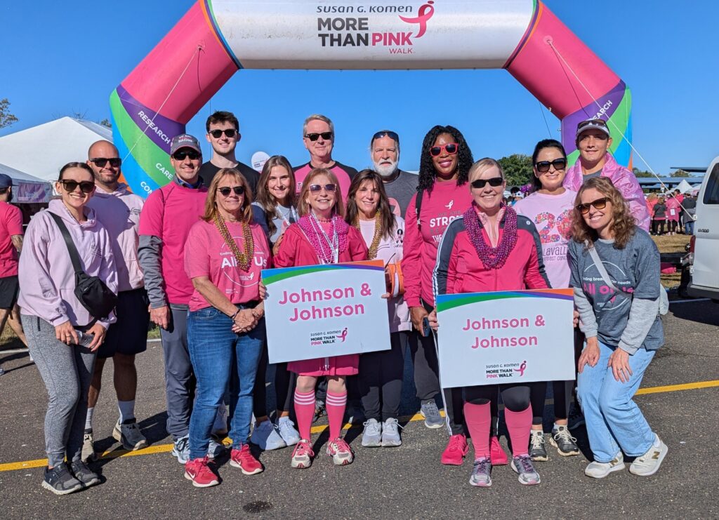 A group of people wearing pink pose together at a charity walk event under a Susan G. Komen More Than Pink arch, holding Johnson & Johnson signs to support breast cancer awareness.