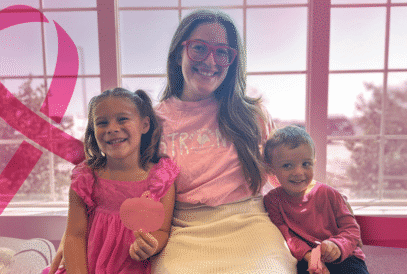 A woman and two children sit together smiling in a sunlit room. The woman wears a "STRONG" shirt and all are dressed in pink. A large pink ribbon symbol is visible on the left.