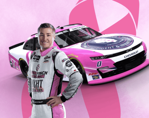 A race car driver in a white and pink suit stands in front of a matching pink and white Chevrolet race car, both featuring racing logos, against a pink backdrop with ribbon designs, highlighting support for National Breast Cancer Awareness Month.