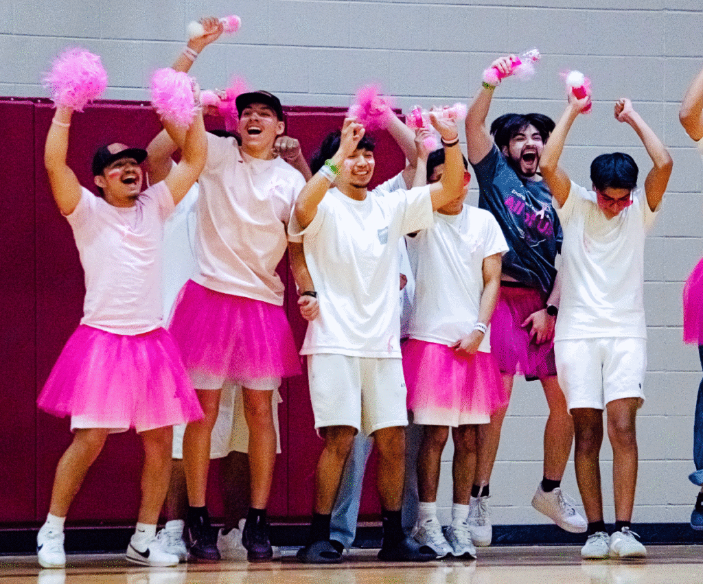 A group of joyful young men wearing pink tutus, pink shirts, and white shorts cheer enthusiastically in a gymnasium, holding pink pom-poms and celebrating together.