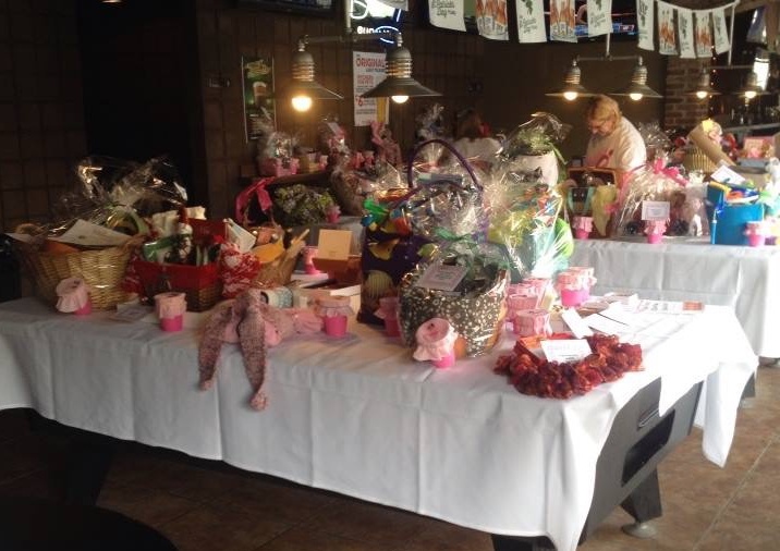 An image of long tables with white tablecloths covered in gift baskets for a breast cancer fundraiser.