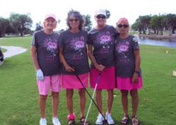 Four smiling women stand together outdoors at a golf breast cancer fundraiser, wearing matching gray shirts with pink floral designs and “I’m Still Standing – Breast Cancer Awareness” printed on them.