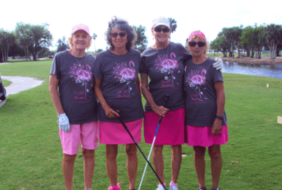 Four smiling women stand together outdoors at a golf breast cancer fundraiser, wearing matching gray shirts with pink floral designs and “I’m Still Standing – Breast Cancer Awareness” printed on them.