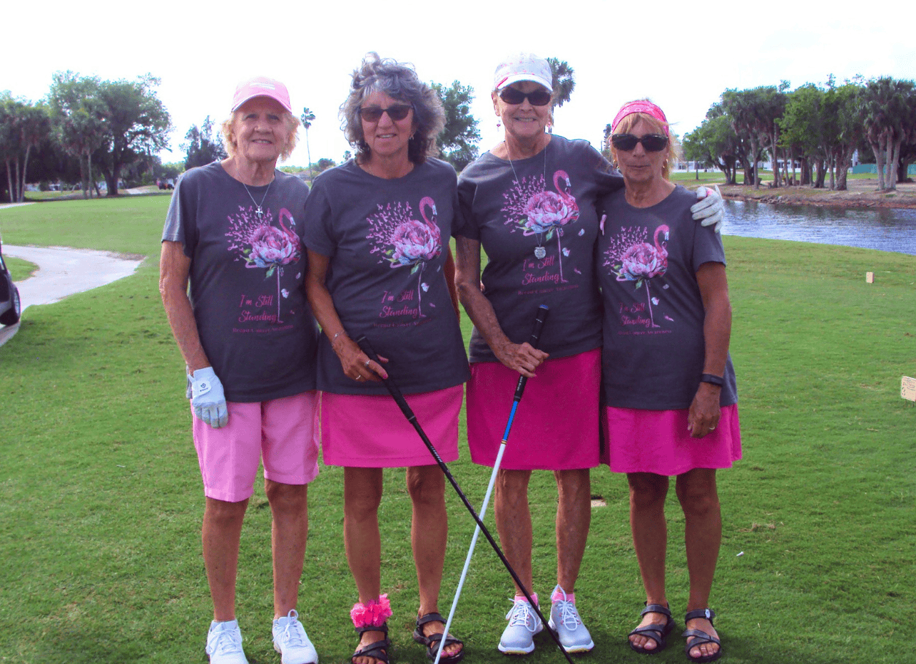 Four smiling women stand together outdoors at a golf breast cancer fundraiser, wearing matching gray shirts with pink floral designs and “I’m Still Standing – Breast Cancer Awareness” printed on them.