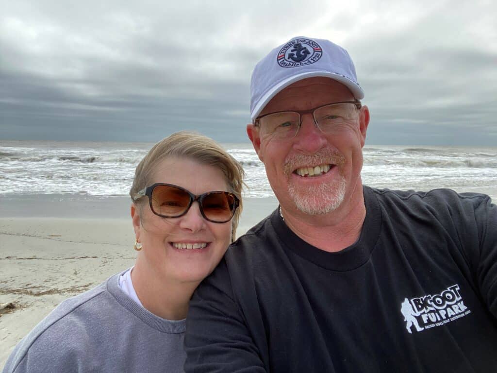 Breast cancer survivor Gretchen and her husband smile on the beach with waves behind them.