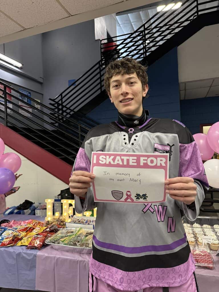 A youth hockey player wearing a breast cancer awareness jersey stands in front of a table with a variety of snacks. He is holding a sign that says, "I Skate for In Memory of my Aunt Mary."