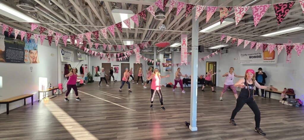 A group of women in athletic wear join a jazzercise breast cancer fundraiser in a bright studio decorated with pink awareness flags and banners. Sunlight streams through windows, and motivational posters are on the walls.