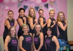 A group of ten women pose together, smiling and flexing their arms in matching black "dance FIGHT CURE" tank tops, in front of a pink, balloon-decorated backdrop at a jazzercise breast cancer fundraiser.
