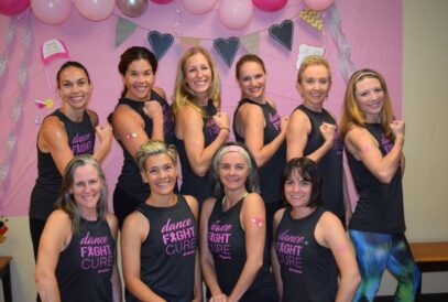 A group of ten women pose together, smiling and flexing their arms in matching black "dance FIGHT CURE" tank tops, in front of a pink, balloon-decorated backdrop at a jazzercise breast cancer fundraiser.