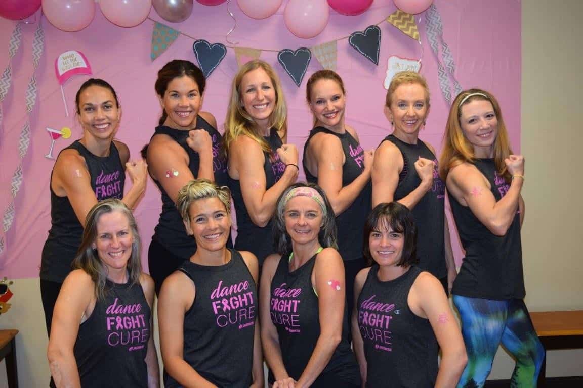 A group of ten women pose together, smiling and flexing their arms in matching black "dance FIGHT CURE" tank tops, in front of a pink, balloon-decorated backdrop at a jazzercise breast cancer fundraiser.