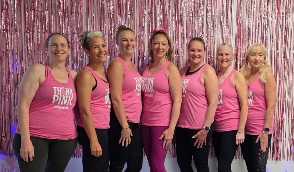 Seven women wearing matching pink “Think Pink” tank tops stand side by side, smiling in front of a pink tinsel curtain at a jazzercise breast cancer fundraiser.