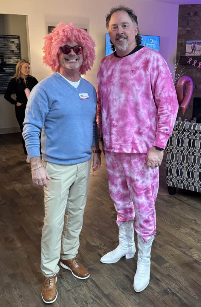 Two men stand indoors, one wearing a pink wig, heart-shaped glasses, blue sweater, and khakis, the other in a pink tie-dye tracksuit and white boots - showcasing their support for Komen as VIPink Ambassador Fundraisers. Both are smiling; a third person is blurred in the background.