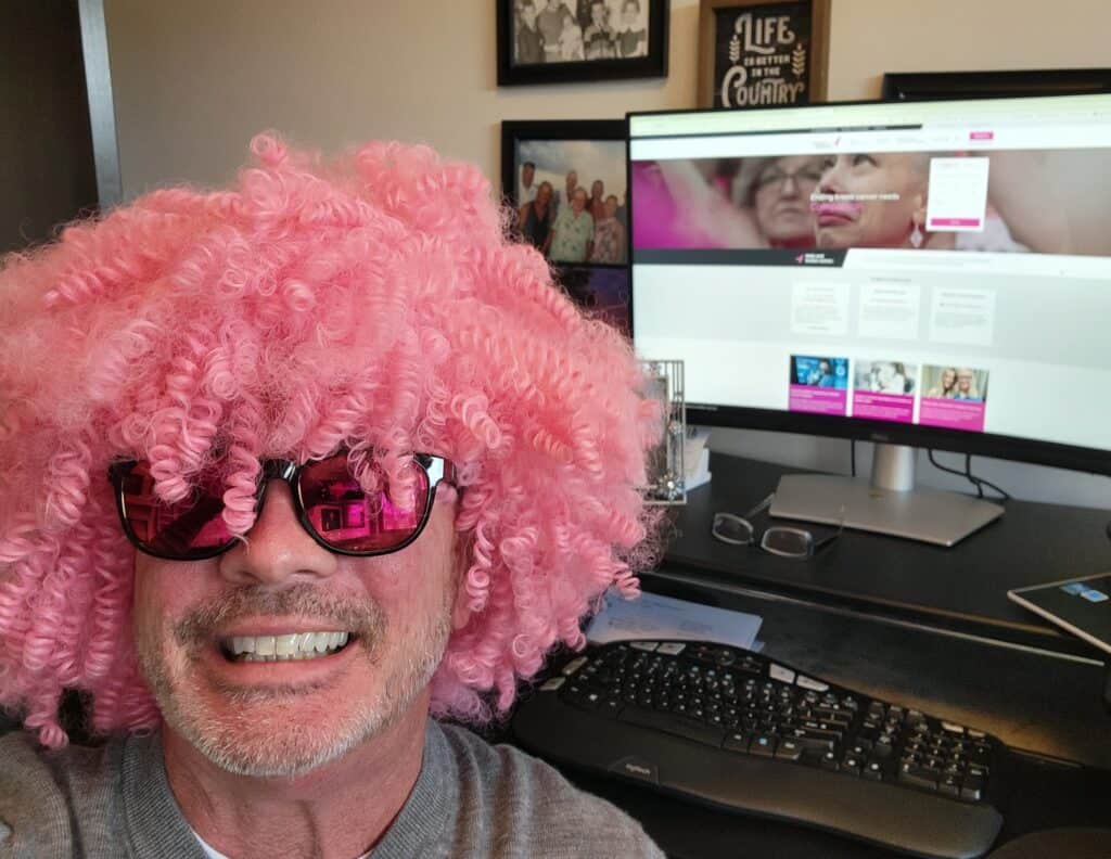 A smiling VIPink ambassador wearing a curly pink wig and sunglasses sits at a desk with a computer. The monitor displays a fundraising website with pink elements, while framed photos and a decorative sign are visible in the background.