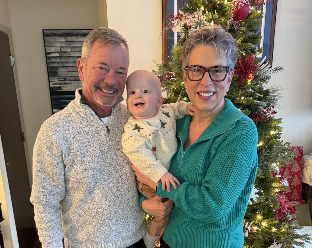 An older man and woman smile and pose with a baby in front of a decorated Christmas tree and wrapped gifts in a cozy living room.