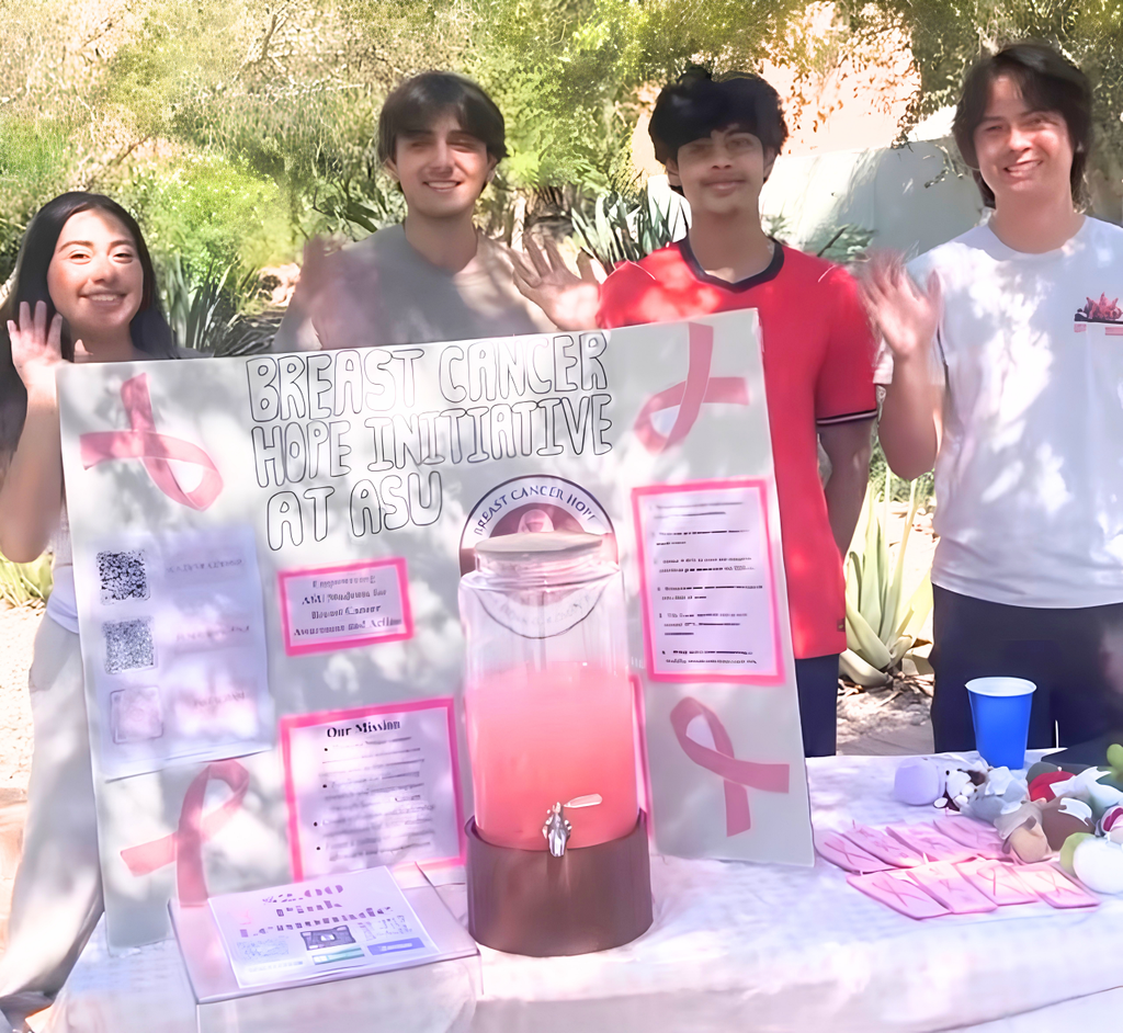 Four students from the breast cancer awareness club stand behind a table with an initiative poster, a pink drink dispenser, and informational flyers, waving at the camera.