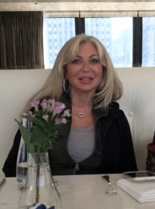 A blonde woman with a heart-shaped necklace sitting at a table with a small vase of flowers.