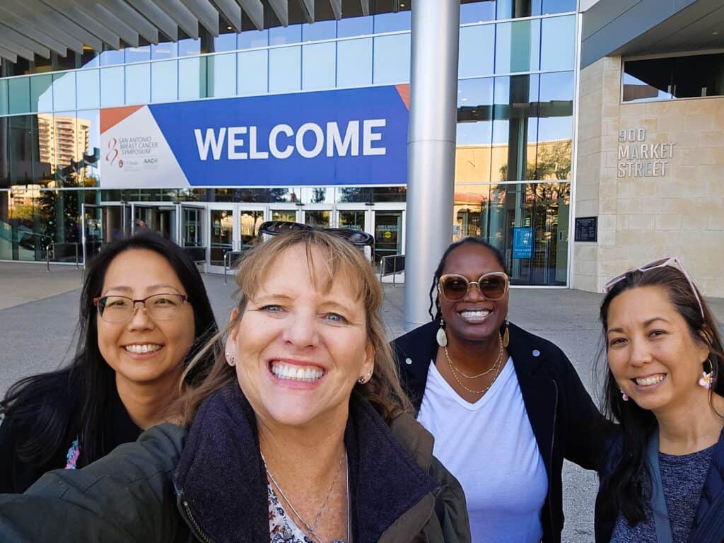 Four patient advocates take a selfie at SABCS, in front of a large sign that reads "Welcome."