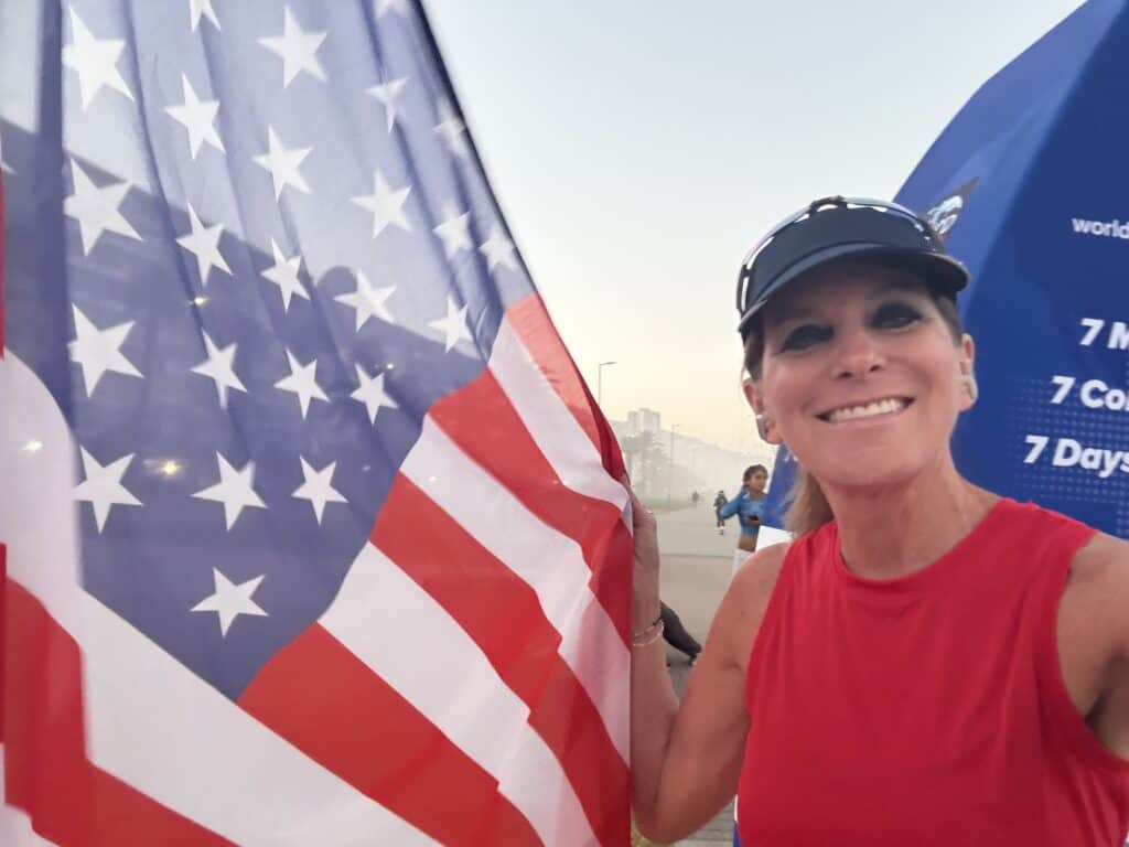 A smiling woman in a red sleeveless top and visor holds a large American flag outdoors. There are buildings and other people in the background, with a blue sky and a slight haze.