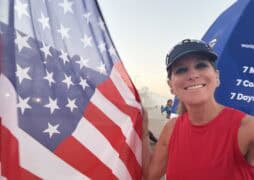 A smiling woman in a red sleeveless top and visor holds a large American flag outdoors. There are buildings and other people in the background, with a blue sky and a slight haze.
