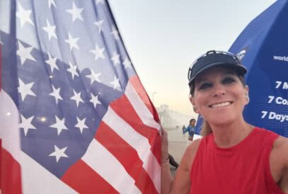 A smiling woman in a red sleeveless top and visor holds a large American flag outdoors. There are buildings and other people in the background, with a blue sky and a slight haze.