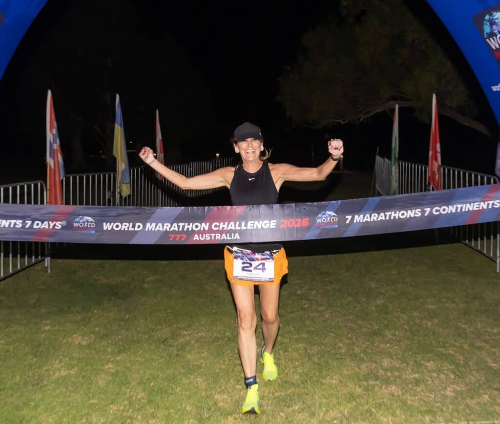 A runner wearing a black tank top and cap crosses the finish line at night, smiling with arms raised, during the World Marathon Challenge 2025 in Australia. The finish banner and country flags are visible behind.