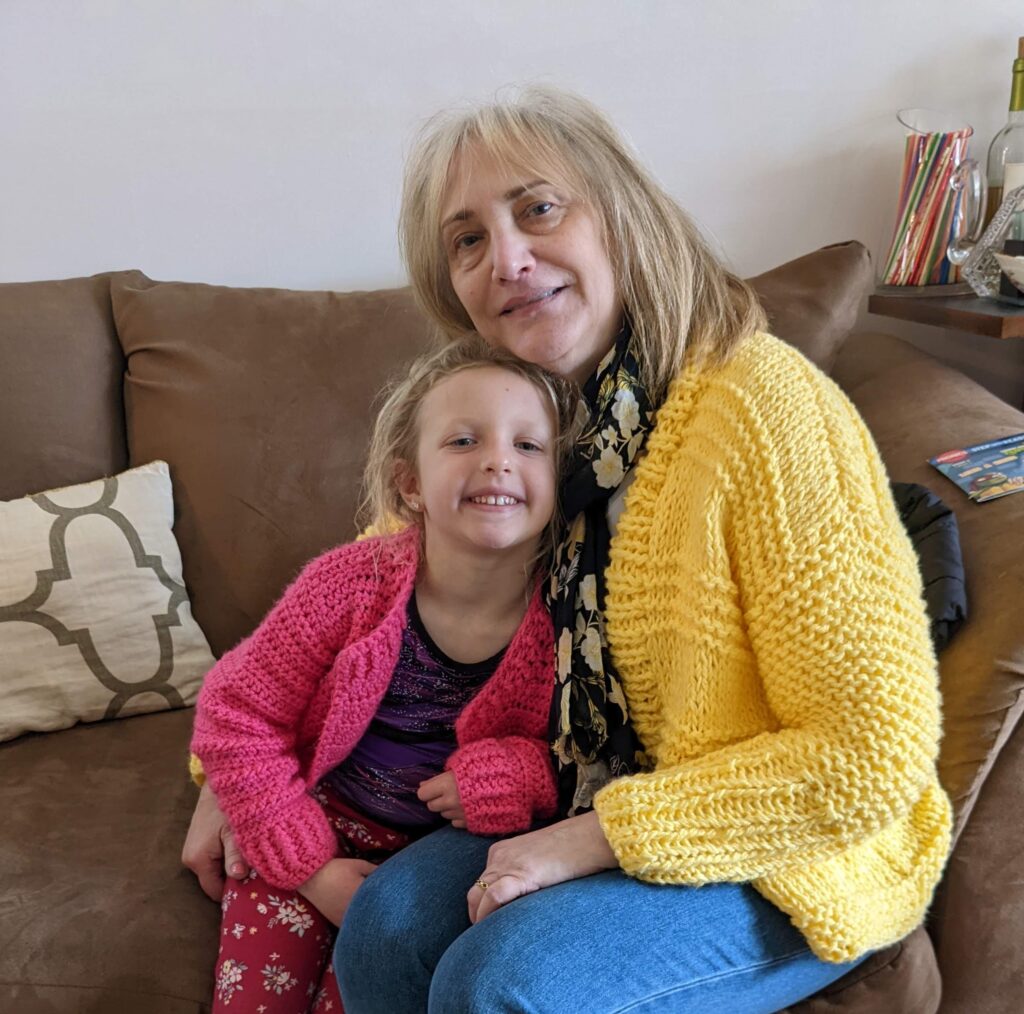 A smiling older woman in a yellow cardigan sits on a couch, hugging a young girl in a pink sweater. They are both looking at the camera, appearing happy and close.