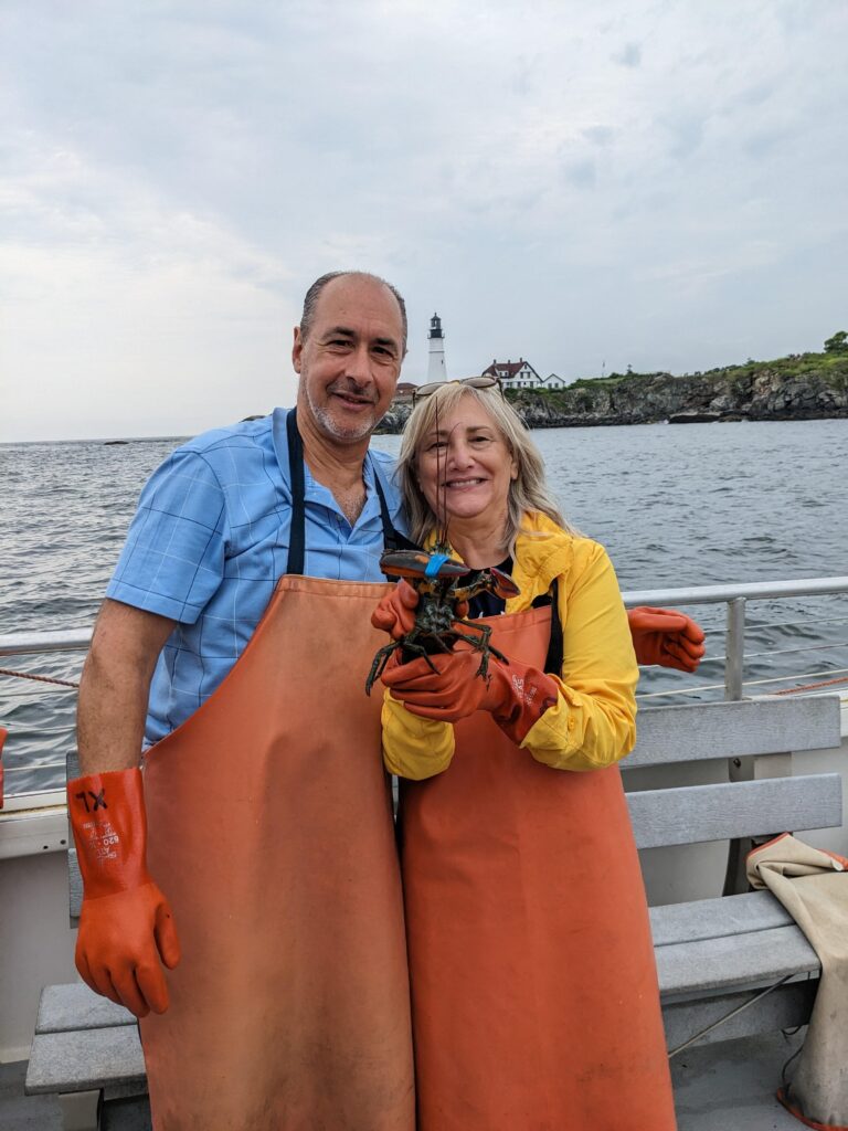 A man and woman in orange aprons and gloves stand on a boat, smiling and holding a lobster. The ocean and a lighthouse on a rocky shore are visible in the background under a cloudy sky.
