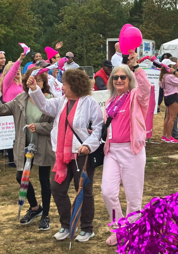 A group of women wearing pink clothing and accessories smile and wave during an outdoor breast cancer awareness event. One woman holds a pink hat, and others carry scarves and umbrellas. Trees and banners are visible in the background.
