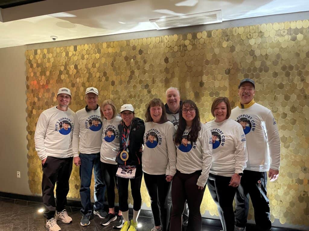A group of nine people wearing matching white “Support Crew” shirts and standing together smiling in front of a gold, circular-patterned wall. One person in the center wears a marathon medal and race bib. They are celebrating the completion of the World Marathon Challenge and the DIY breast cancer fundraiser.