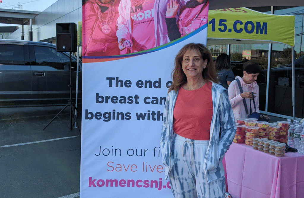 A woman in a blue-and-white tie-dye outfit stands in front of a breast cancer awareness banner at an outdoor event, with a table of snacks and drinks, and another woman organizing items in the background.