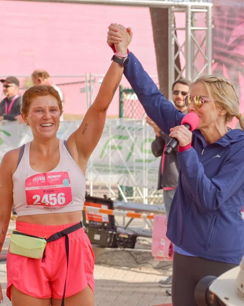 Kayla has crossed a finish line at a race to raise awareness for breast cancer. She is in pink and smiling, her hair growing in.