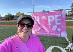A woman in sunglasses, a cap, and a bright pink breast cancer awareness shirt smiles in front of a "HOPE" banner on a sports field. The banner promotes breast cancer awareness with pink ribbons.