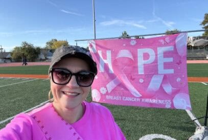 A woman in sunglasses, a cap, and a bright pink breast cancer awareness shirt smiles in front of a "HOPE" banner on a sports field. The banner promotes breast cancer awareness with pink ribbons.
