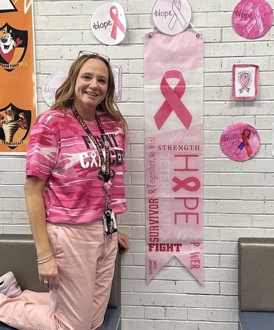 A woman in a pink "Fight Cancer" shirt stands by a wall decorated with pink ribbons and words like "hope" and "strength," promoting breast cancer awareness. She is smiling and wearing an ID badge.