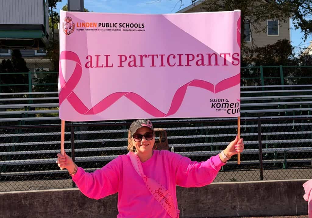 A woman in a pink shirt and cap holds a sign during a school breast cancer walk, reading “Linden Public Schools, all participants, Susan G. Komen for the Cure,” with pink ribbon graphics, while sitting in front of empty bleachers outdoors.